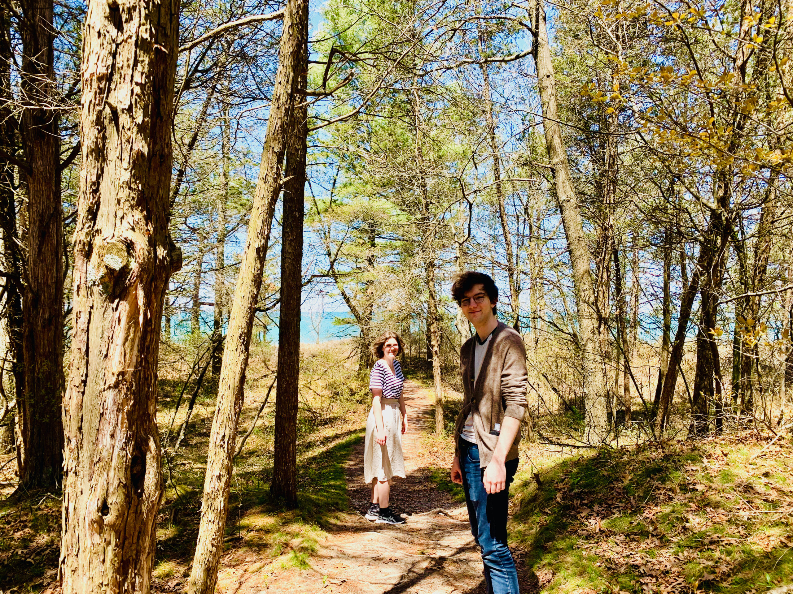 Two teenagers in dappled sunlight on a path in the woods with water in the distance.
