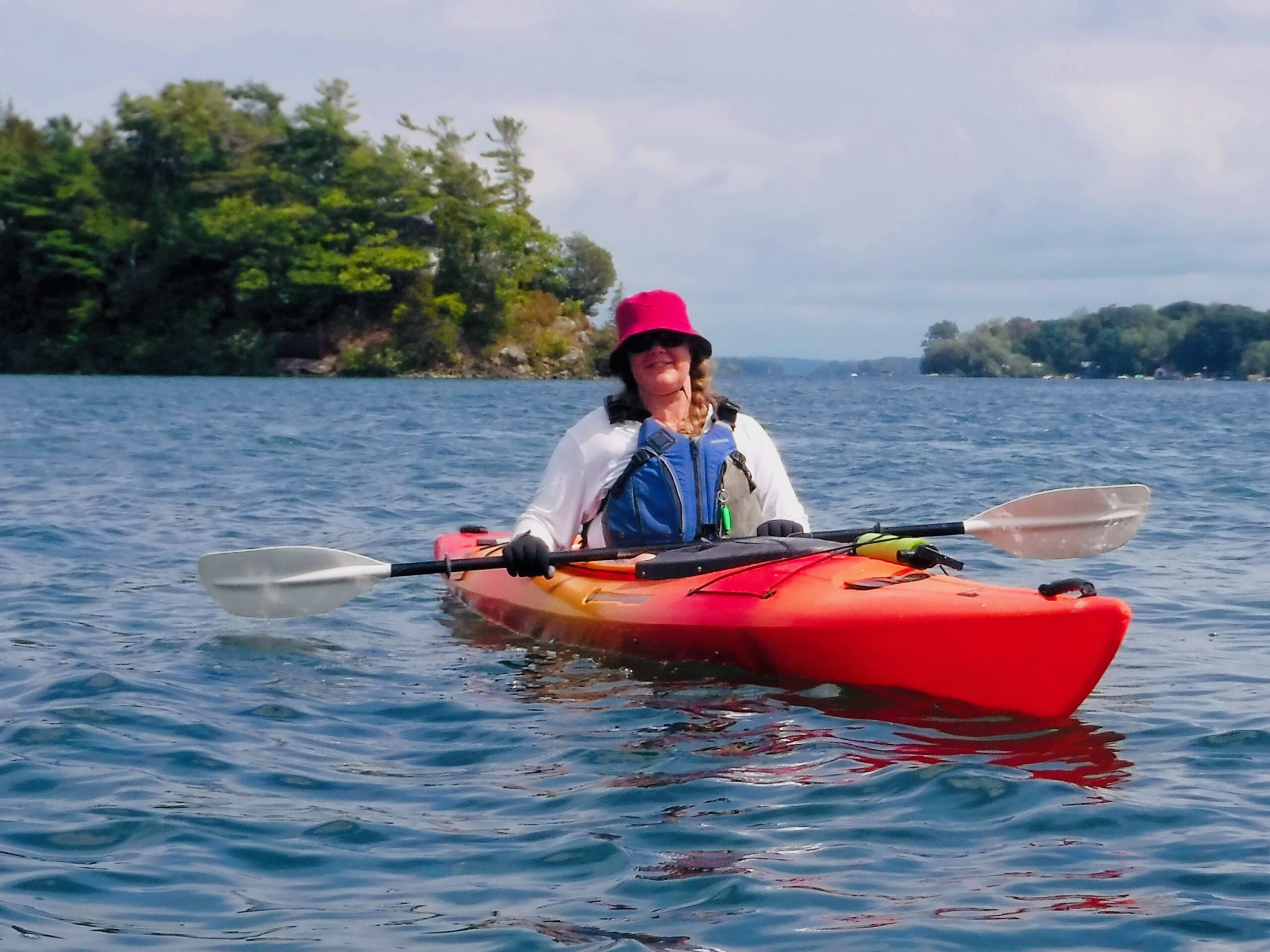 a woman in a red kayak wearing a sun hat and sunglasses (and pfd) resting on the water with islands behind