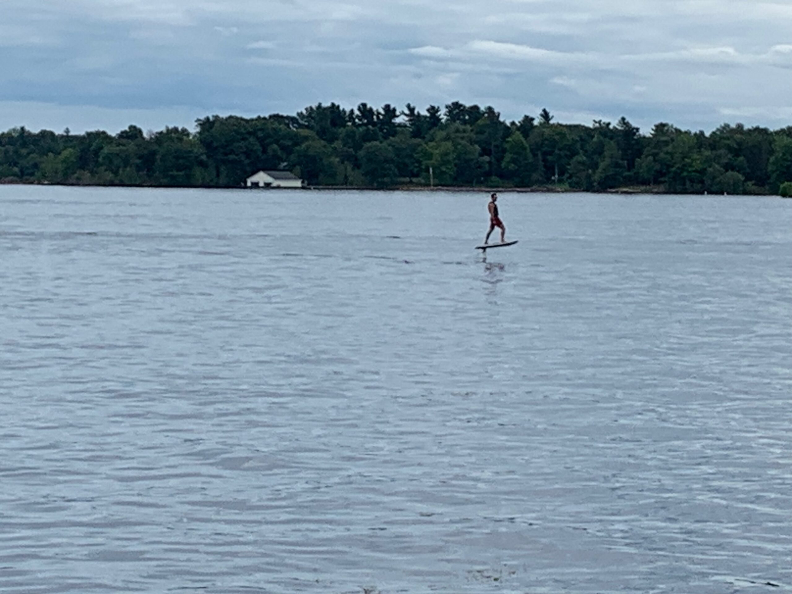 man on a hoverboard (?) on the water with a shoreline behind him