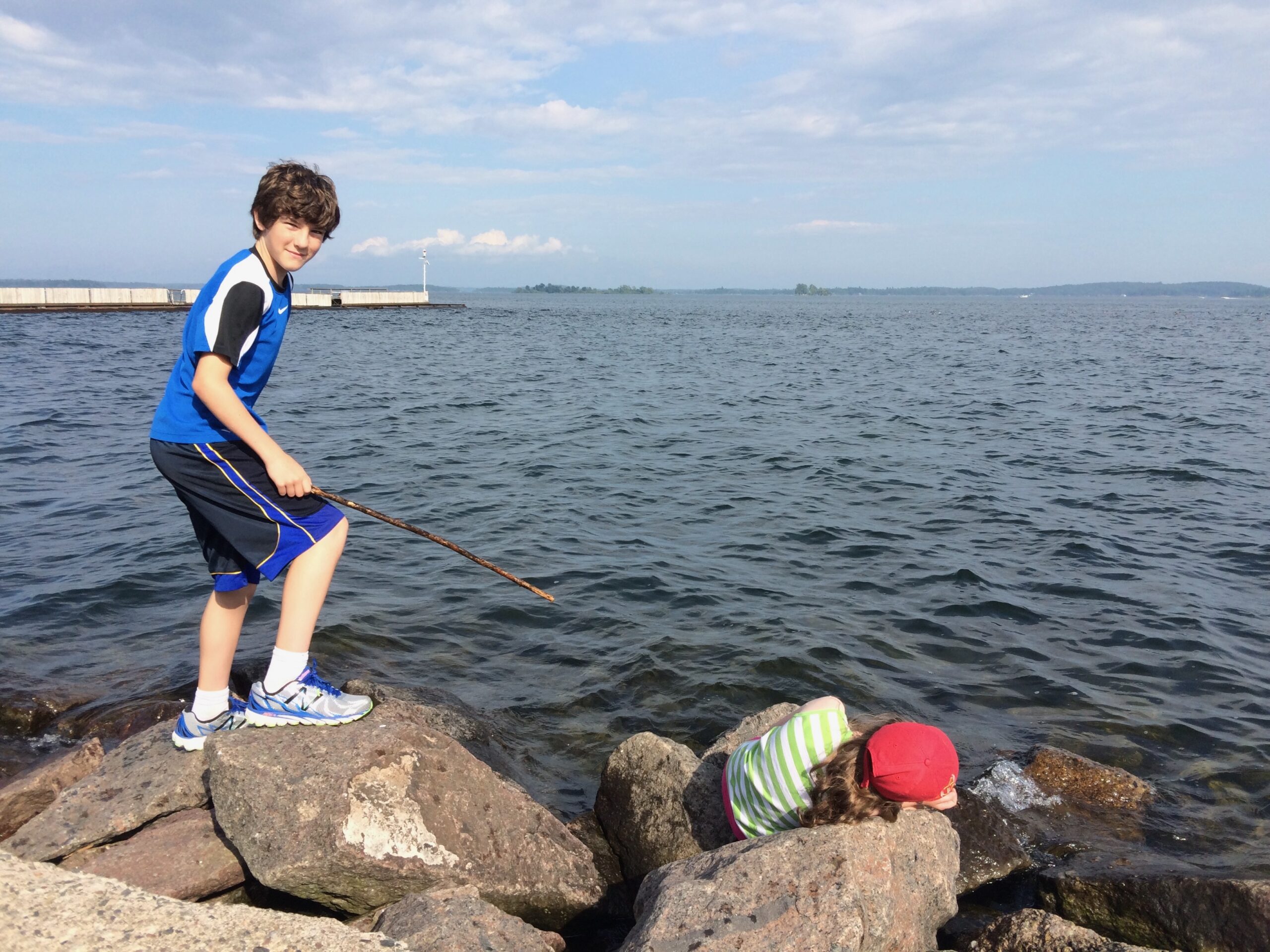 10-year-old boy standing on rocks by water with a stick in his hand and a smile, with a younger girl lying on a rock nearby looking out at the water.