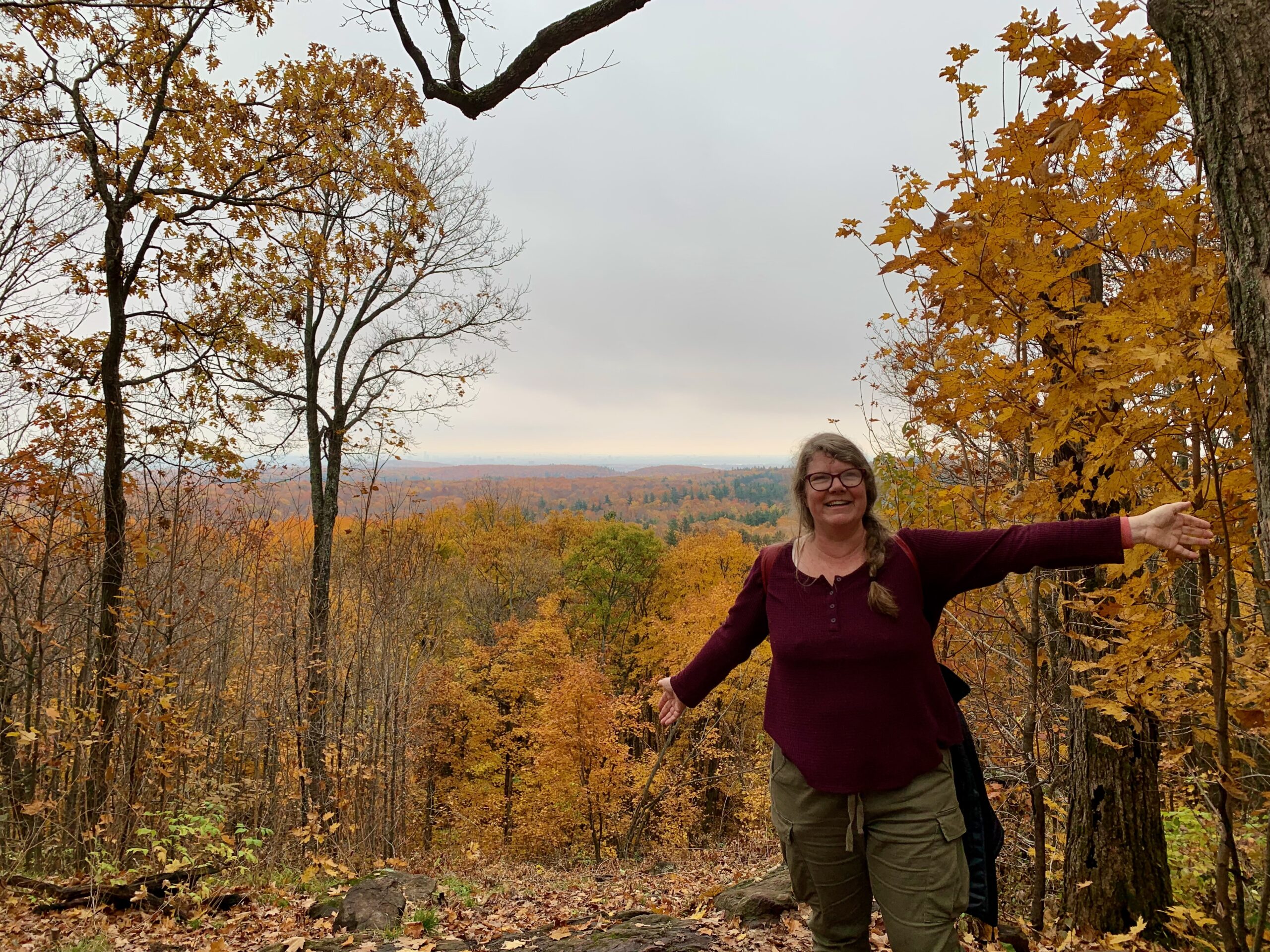 Me standing with arms spread at the top of a hill in the woods, surrounded by fall forest colours.
