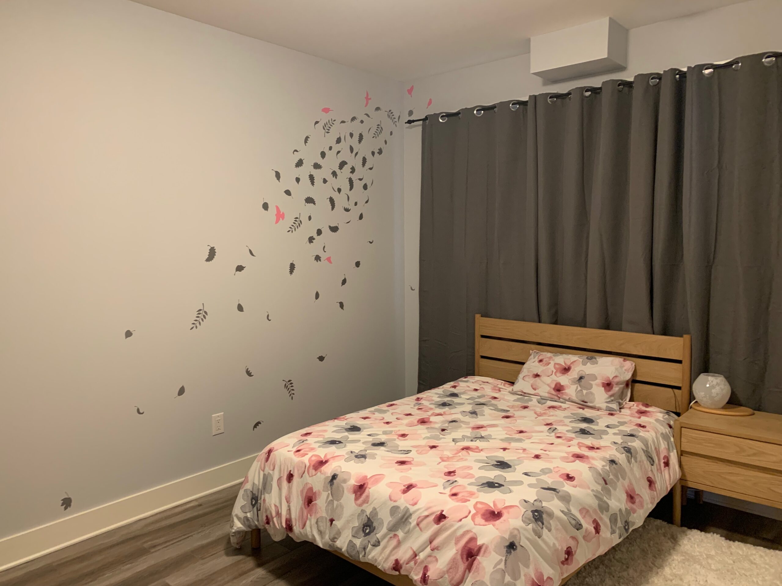 A bedroom with grey curtains, decorations on the wall and a pink and grey bedspread on the bed.