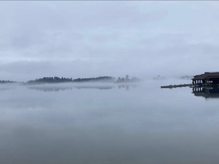 Mist on water smooth as glass with treed islands in the distance and a dock and boat house nearby.